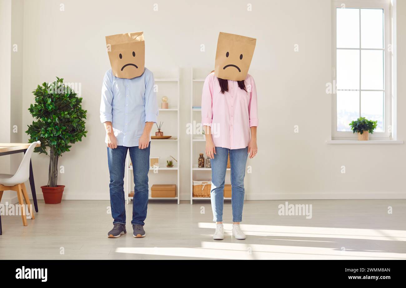 Faceless man and woman standing in the living room at home with paper bags on head with upset faces Stock Photo
