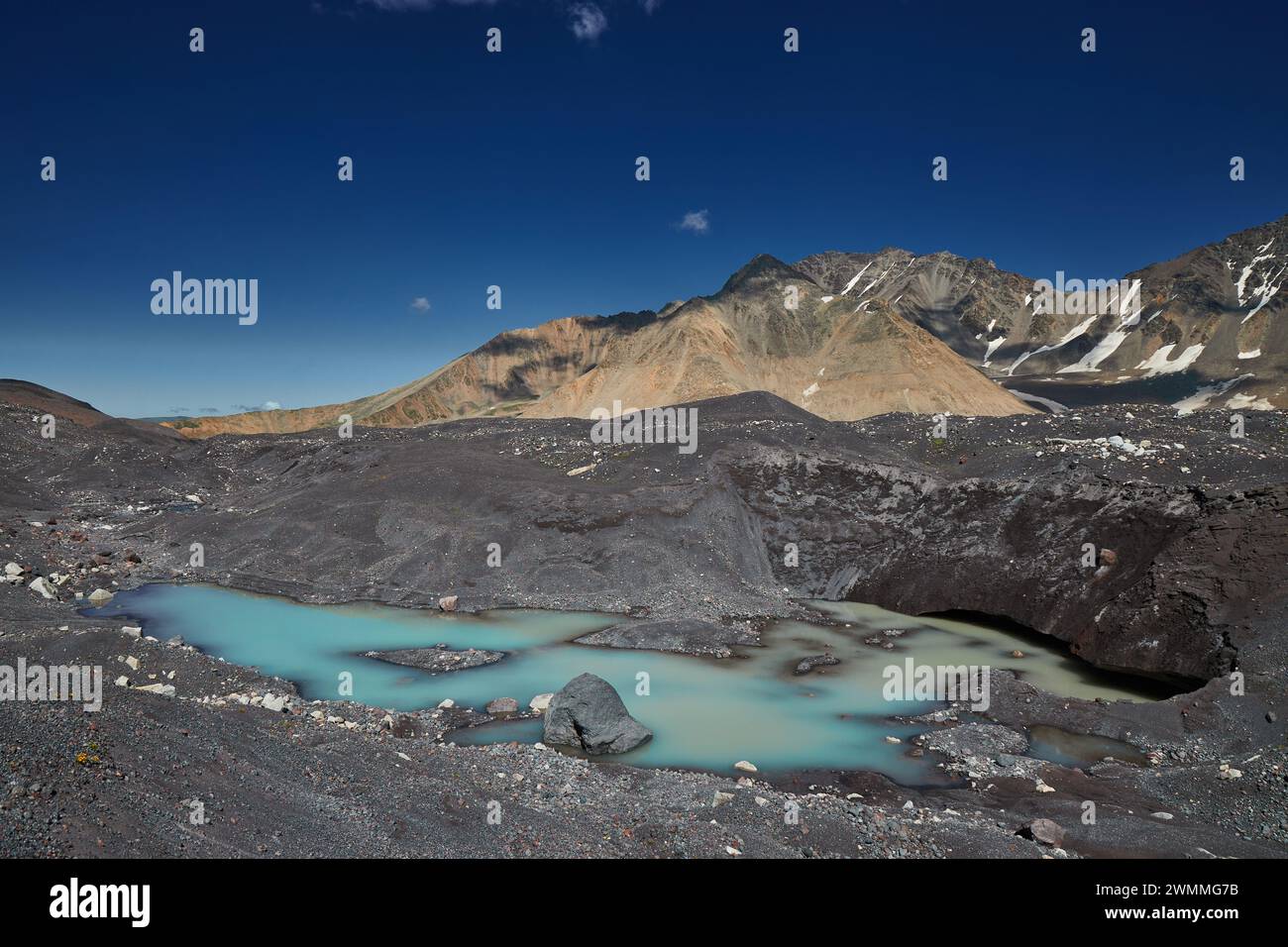 Turquoise glacial lake pool nestles among dark moraines under a stark mountain range and blue sky Stock Photo