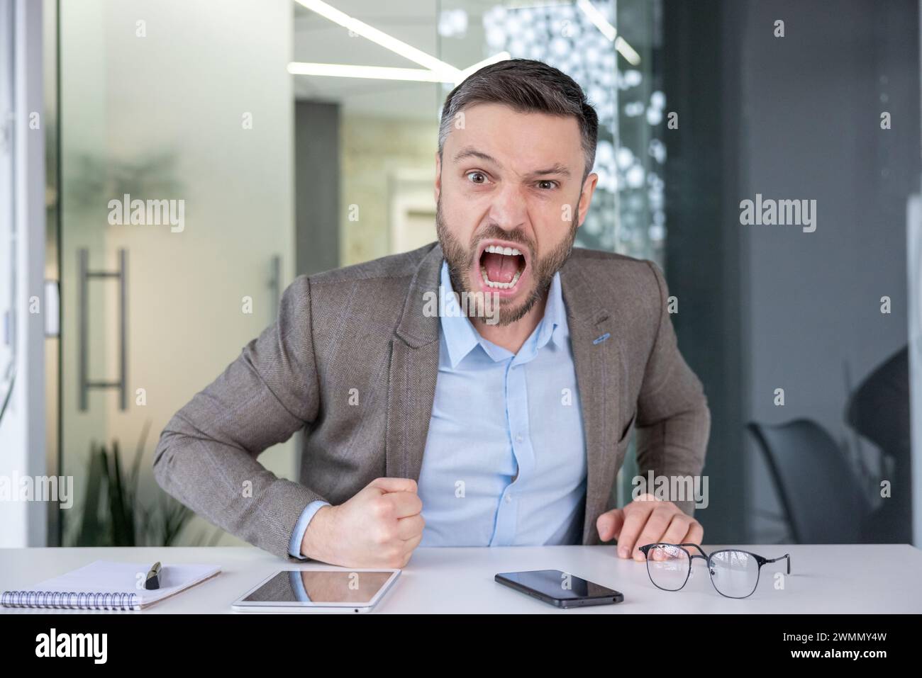 An angry male office worker screaming with clenched fists, showing strong emotion and workplace stress against a corporate interior background. Stock Photo