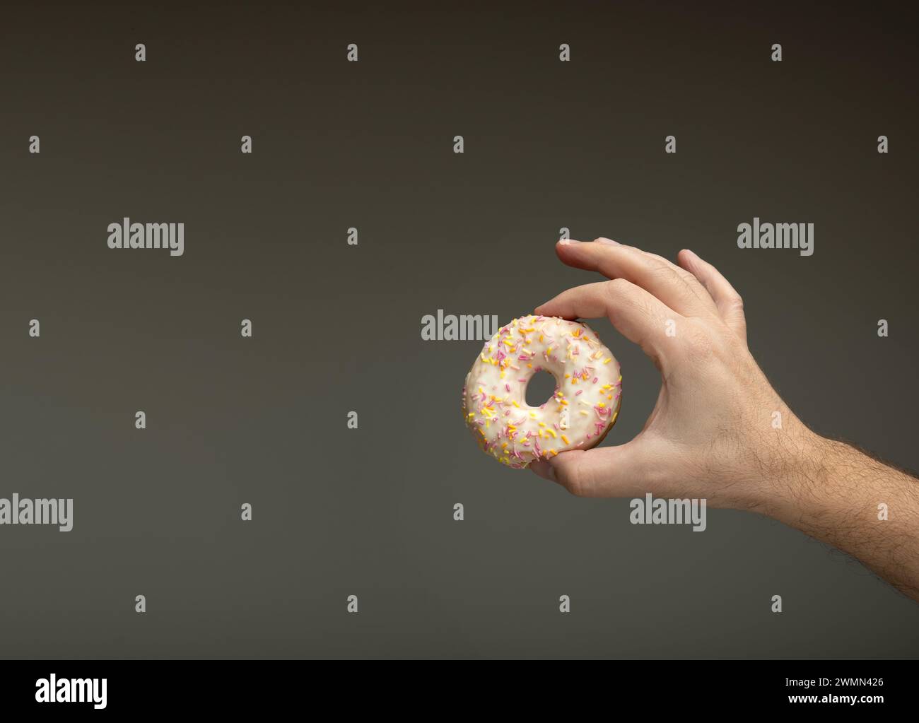 Round donut with frosting and colored sprinkles held between Caucasian man's fingers. Isolated on gray Stock Photo