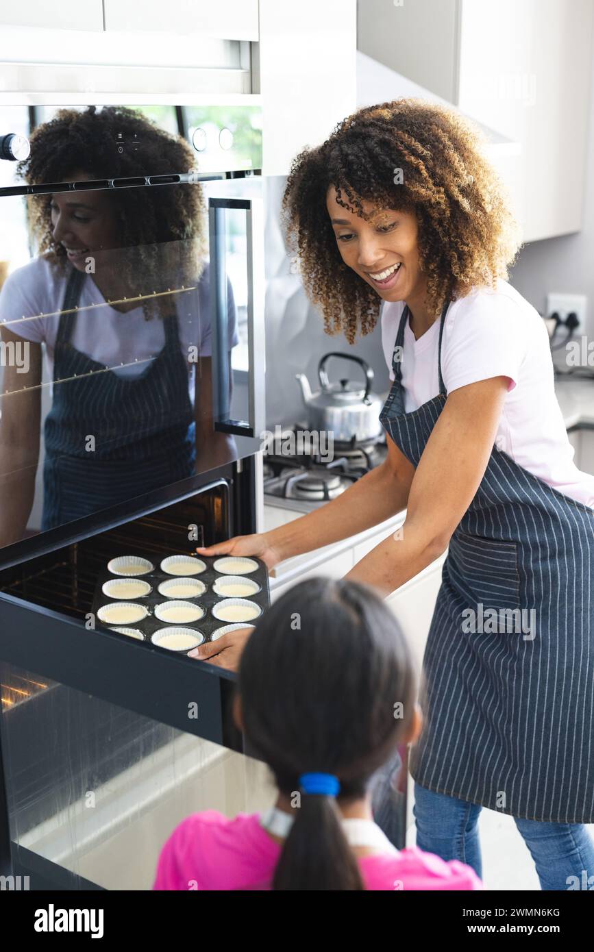 Biracial mother bakes with a biracial daughter at home for her daughter. They share a joyful moment preparing cupcakes in a modern kitchen. Stock Photo