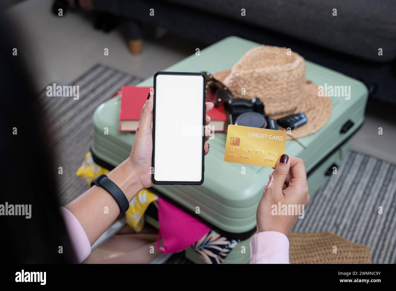 Woman's hand taking a credit card while using a phone, white background with a suitcase in the background. Stock Photo