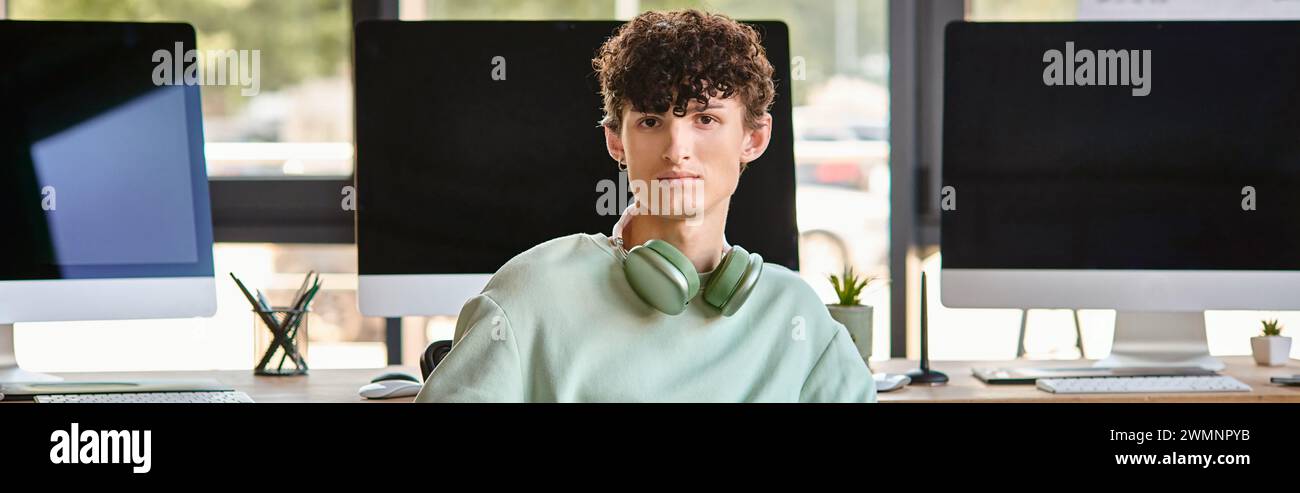 curly haired young man with headphones sitting near computer monitors, post production banner Stock Photo