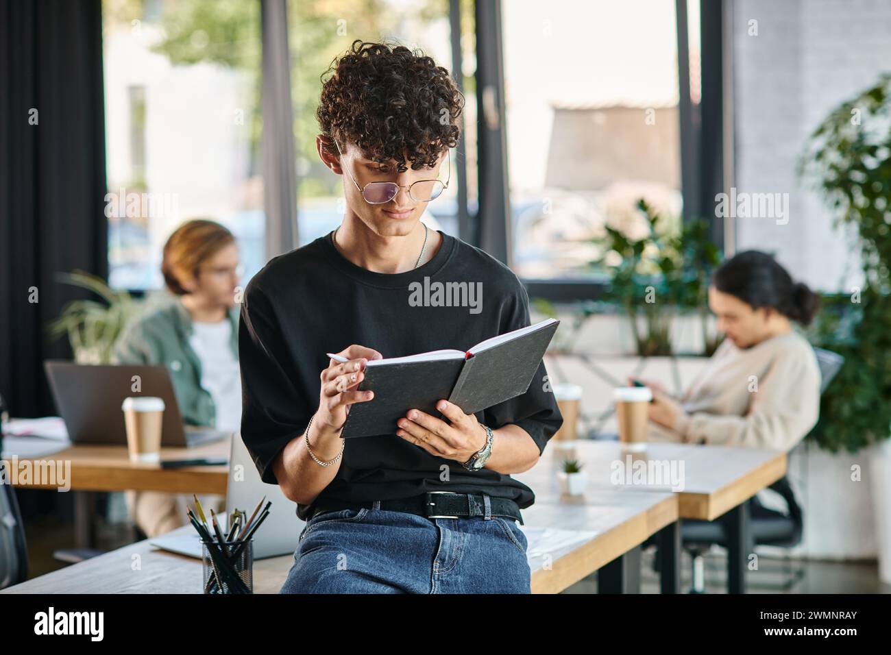 Curly-haired man with glasses taking notes in a dynamic office space, planning startup Stock Photo