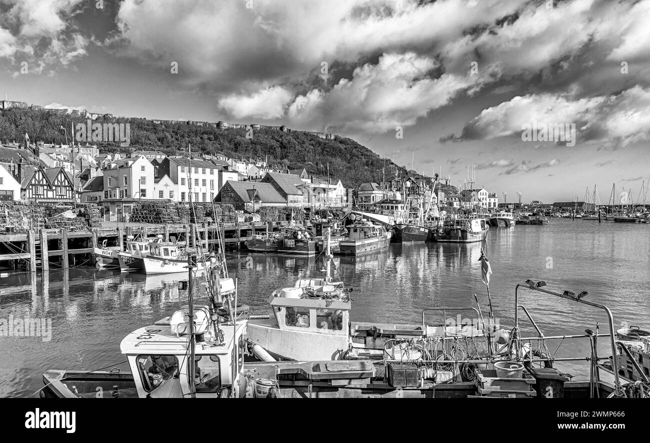 Fishing boats are moored alongside a wharf which is stacked with lobster pots. Buildings line the waterfront and an ancient castle wall is above. Stock Photo