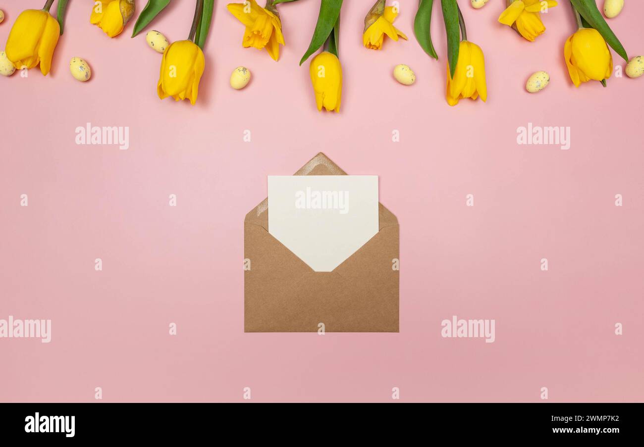 Top view of yellow tulips, daffodils, easter eggs on pink background. Kraft envelope, white card. Spring easter composition, flat lay, copy space. Stock Photo