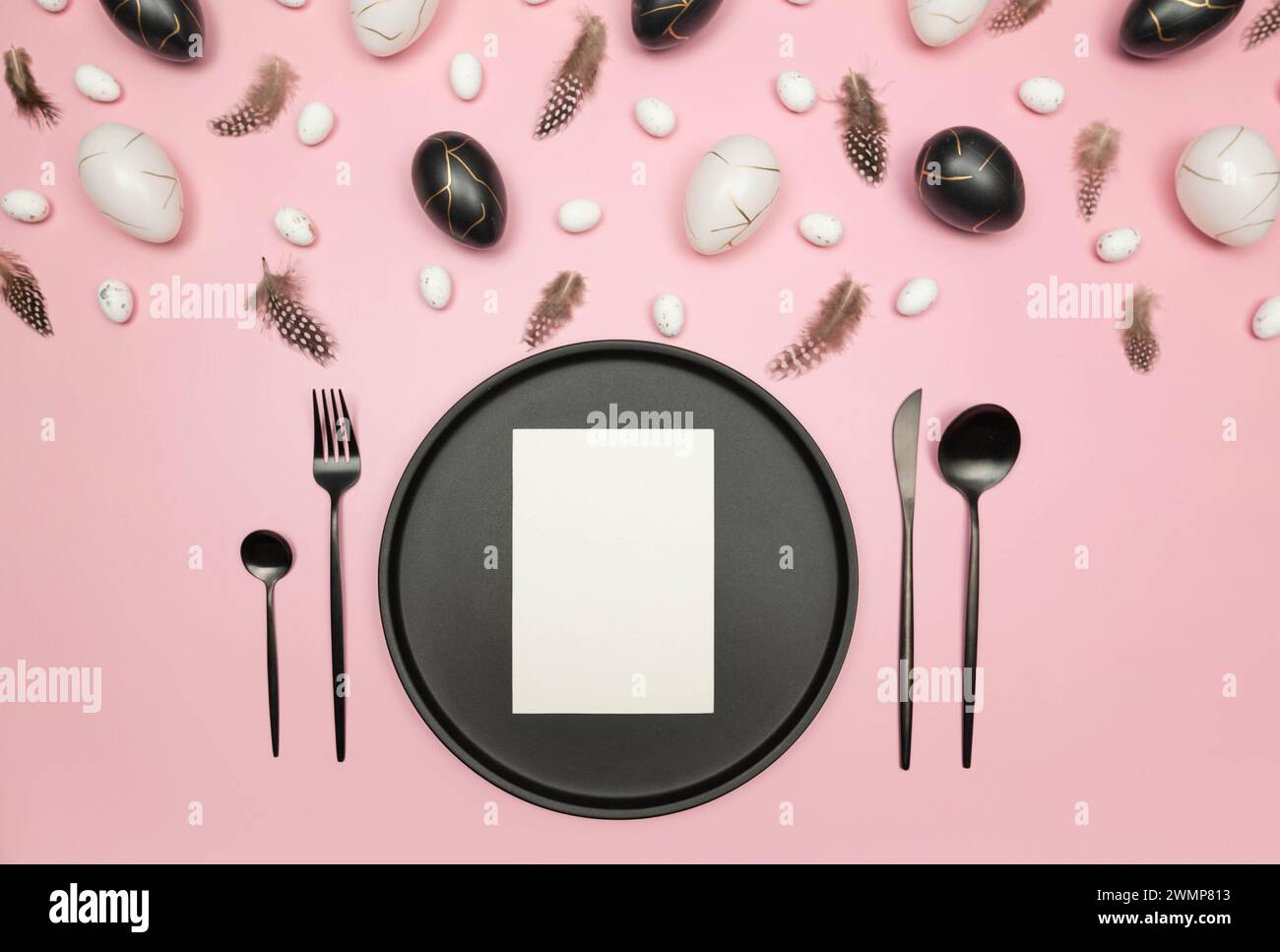 Top view of easter eggs, feathers on pink background. Table setting, black plate and cutlery, white card. Creative easter composition, spring. Stock Photo