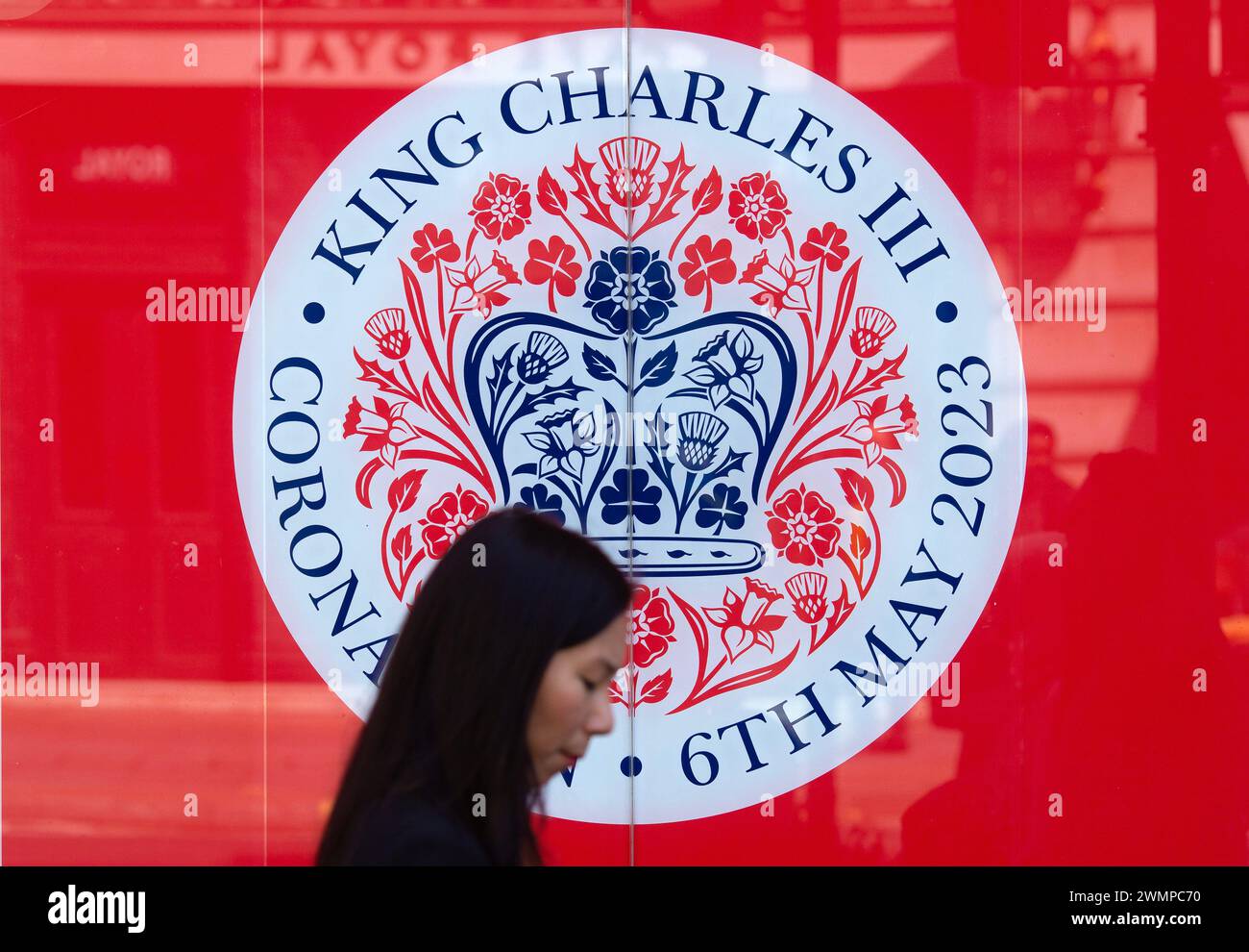 People walk past the coronation emblem as London prepares for the coronation of King Charles III. Stock Photo
