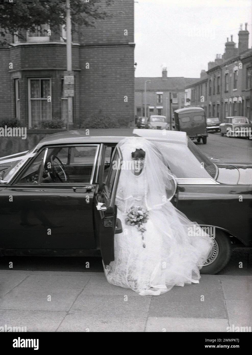 1960s, historical, outside in an urban street, a bride in her wedding dress and veil, getting out of the back of her wedding car, a Ford Zephyr MK3 of the era, England, UK. Stock Photo
