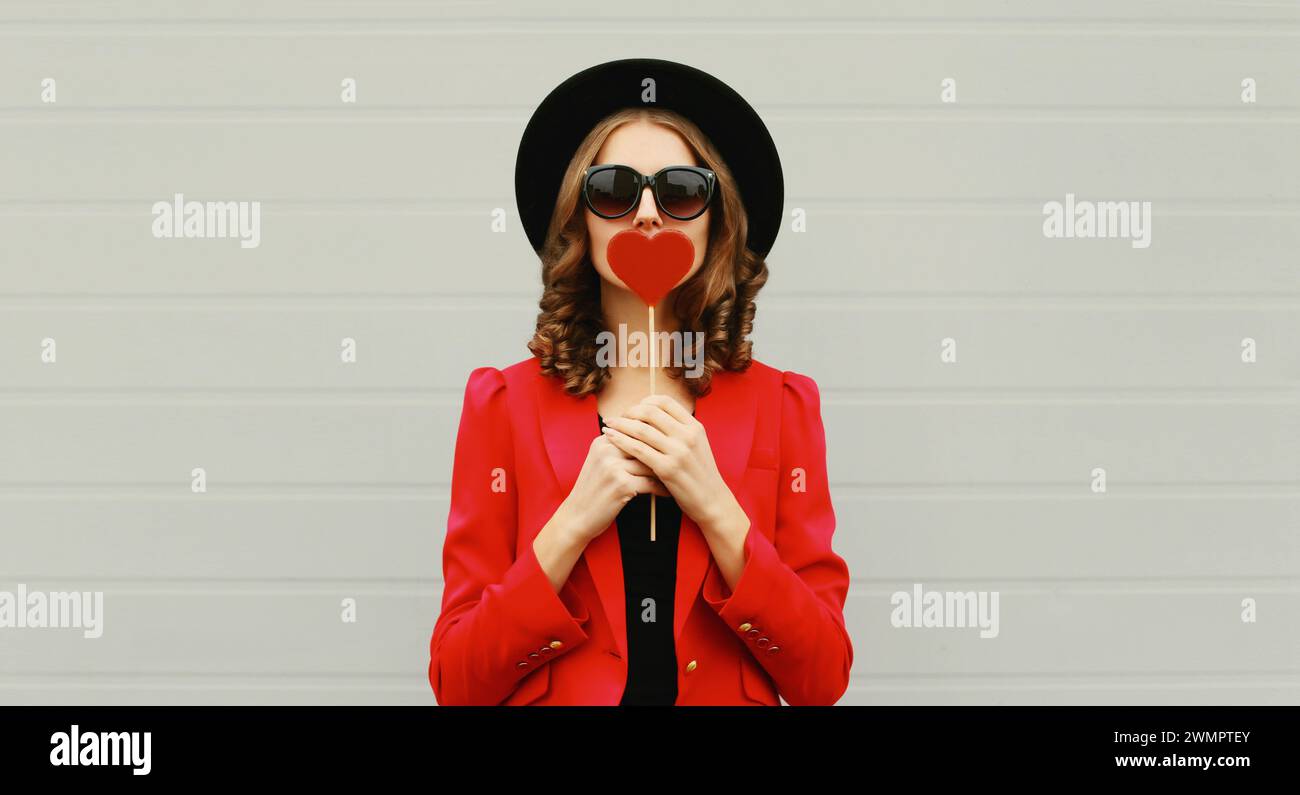 Beautiful stylish woman blowing kiss with sweet red heart shaped lollipop on stick in black round hat Stock Photo