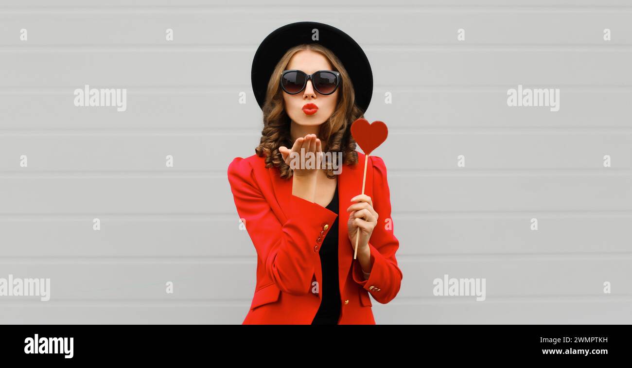Beautiful stylish woman blowing kiss with sweet red heart shaped lollipop on stick in black round hat Stock Photo