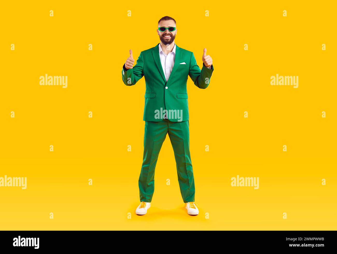 Happy man in green suit standing on yellow background, showing thumbs up and smiling Stock Photo