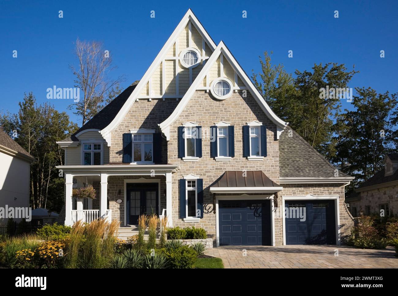 Luxurious Victorian style light brown and tan cut stone home with blue trim, two car garage, landscaped front yard and paving stone driveway in summer. Stock Photo