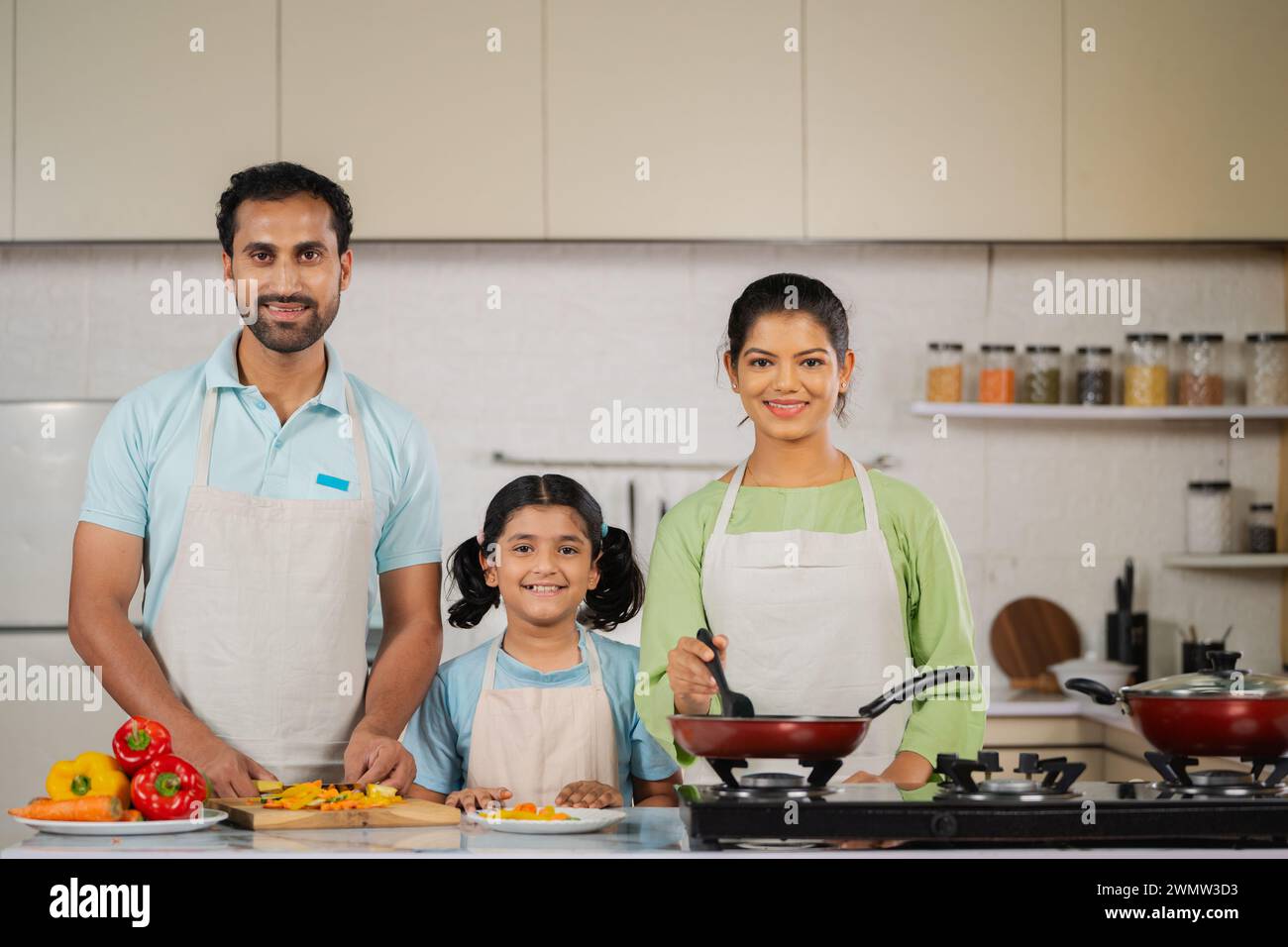 Group of Indian Parents with daughter looking at camera while busy cooking together at kitchen - concept of family bonding, weekend holidays and Stock Photo