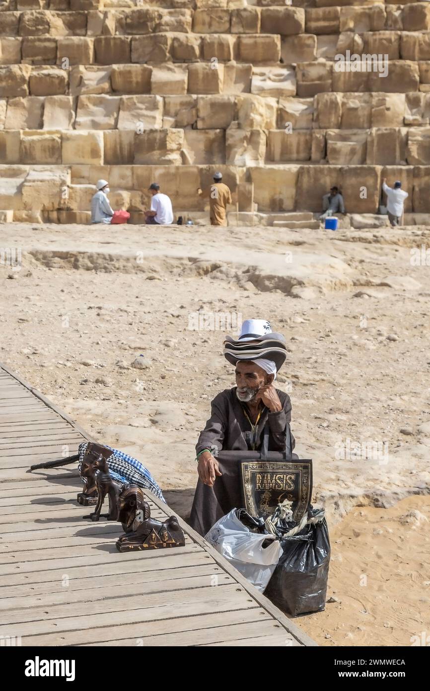 Giza Necropolis, Egypt - April 26, 2022: Old Bedouin man dressed in traditional clothes, sell specific souvenirs in front of the Giza Necropolis pyram Stock Photo