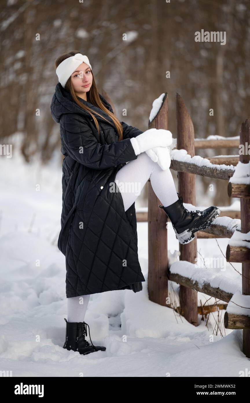 Portrait of a girl in winter clothes with white gloves and a white headband near a wooden fence and forest. Stock Photo