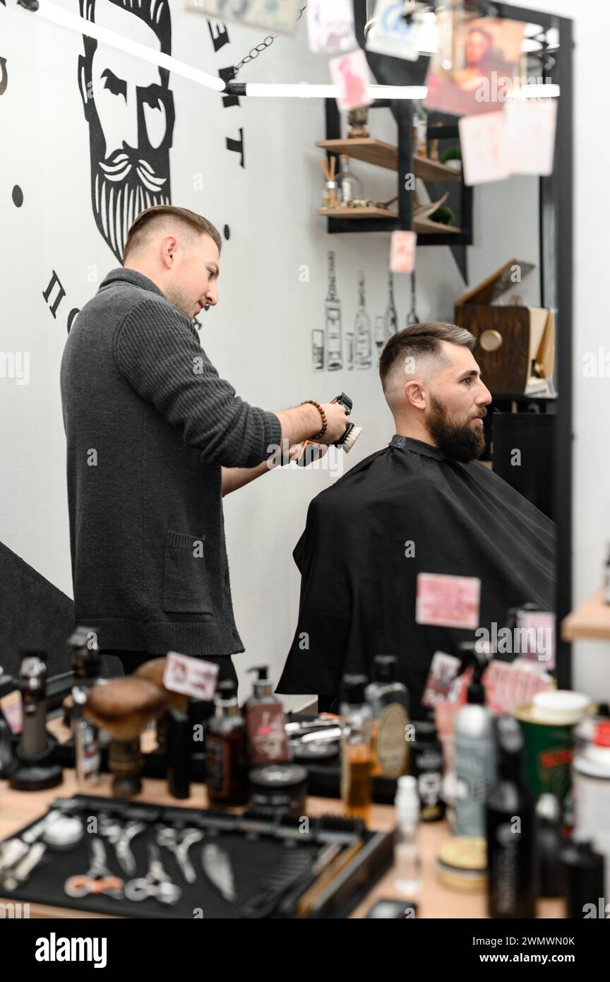 A barber cuts a man with a beard in a barber shop. Short haircut of the client with a clipper. Stock Photo