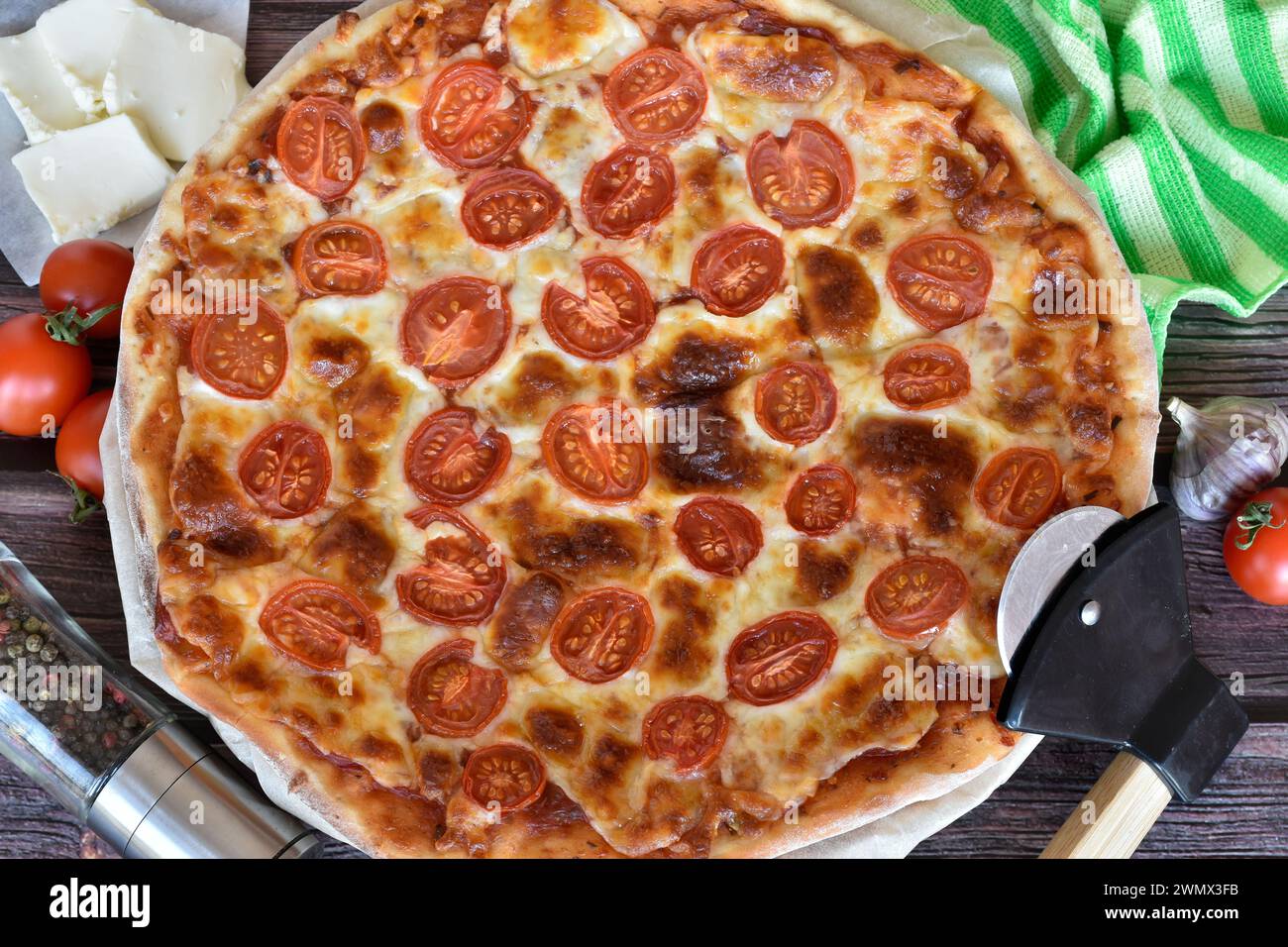 Fresh baked homemade pizza Margherita served on rustic wooden table. Cherry tomatoes, mozzarella cheese. Pizza cutter, pepper mill, garlic, napkin. Stock Photo