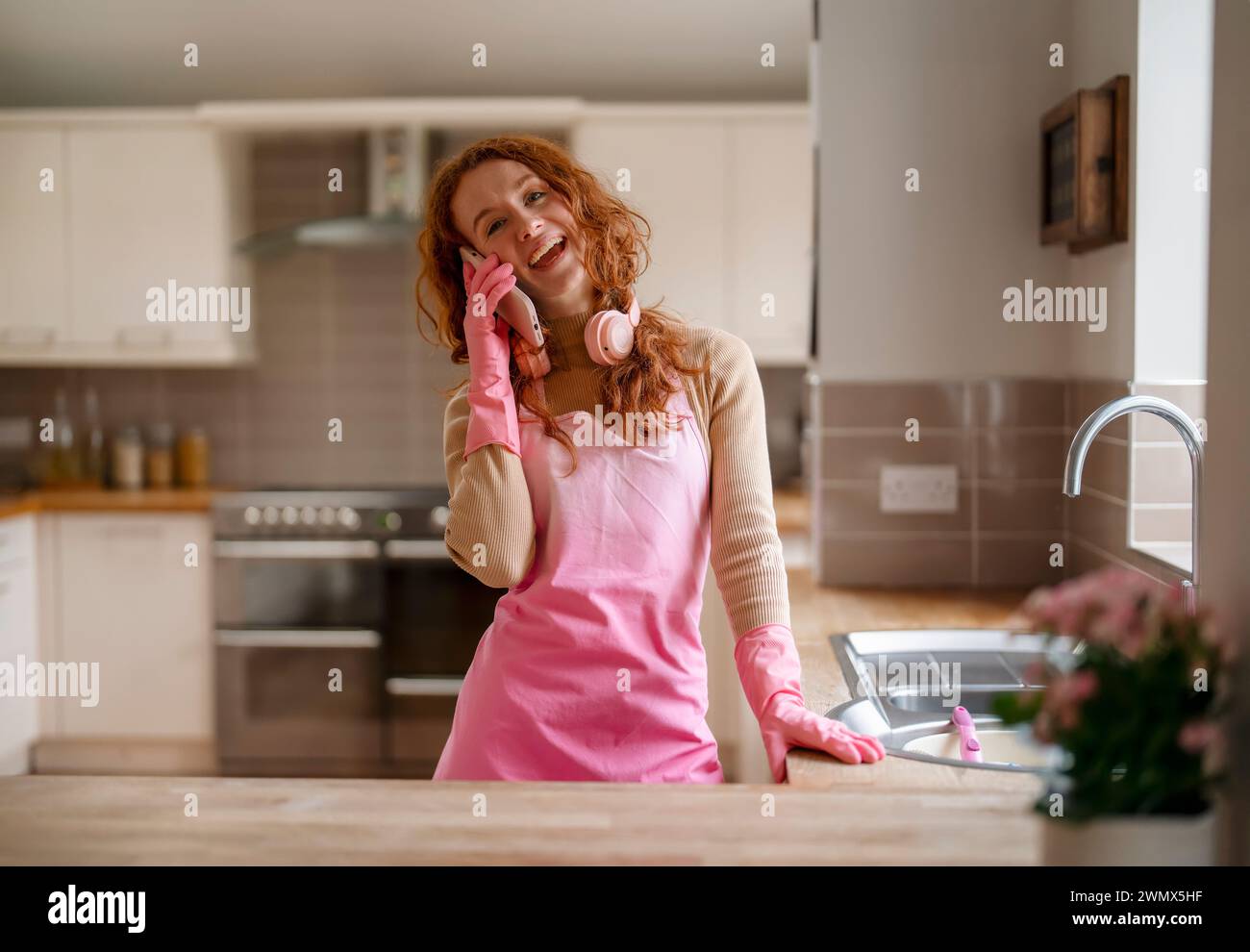 Redhead woman washing dishes in the kitchen and listening to music with wireless headphones, using a mobile phone with domestic gloves, enjoying makin Stock Photo