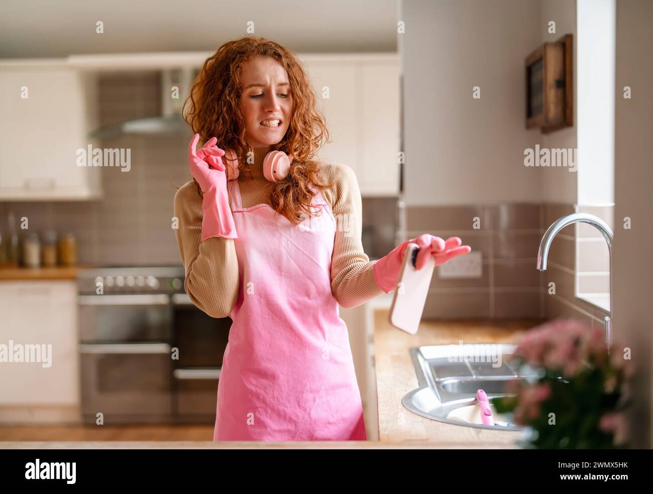 Redhead woman washing dishes in the kitchen and listening to music with wireless headphones, trying to use a mobile phone with domestic gloves, enjoyi Stock Photo