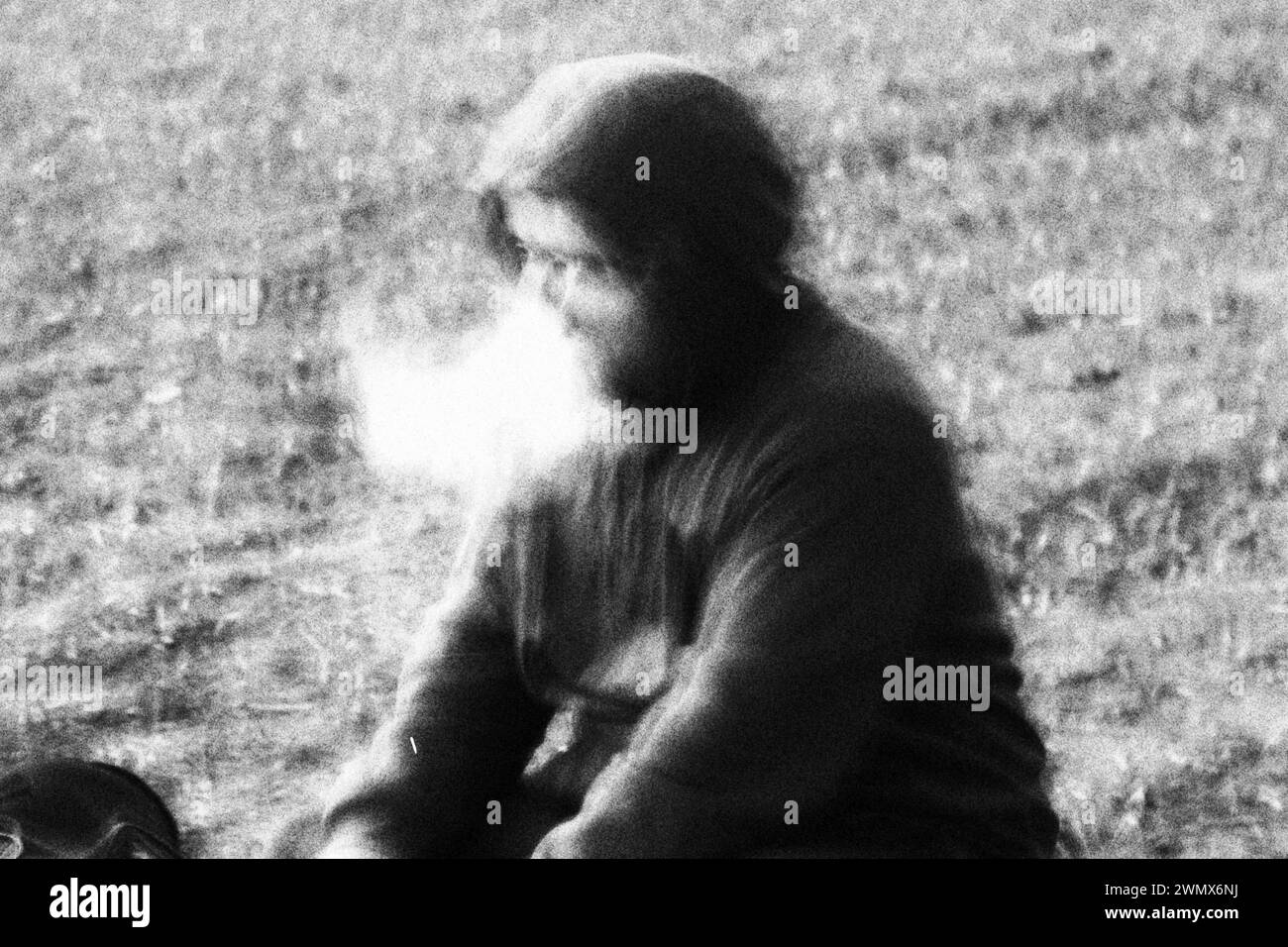 In a scene shrouded in mystery, a person wearing a hoodie emerges from behind a dense cloud of smoke, captured in striking black and white with a nois Stock Photo