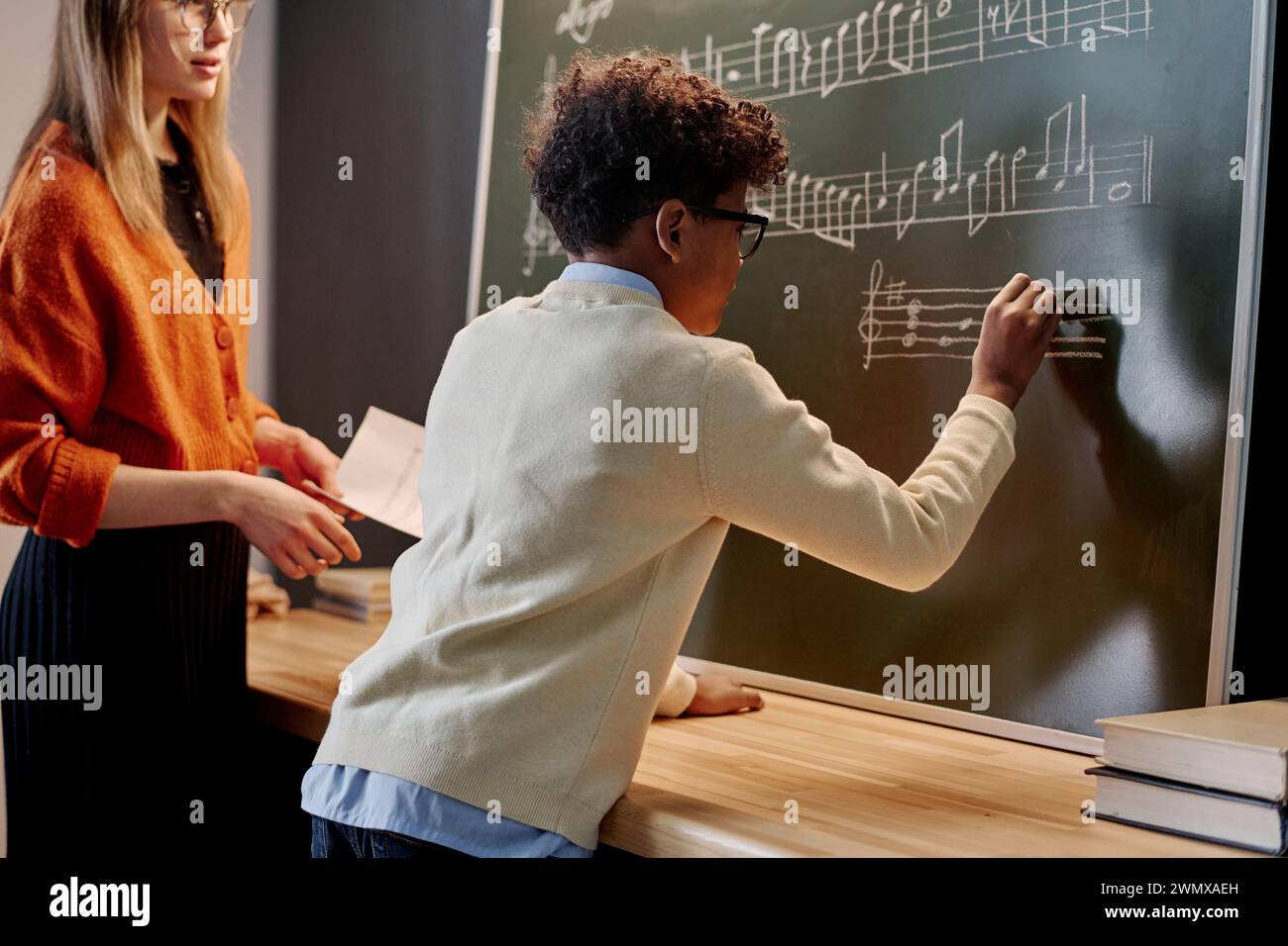 African American boy writing notes blackboard during music class, his Caucasian female teacher standing next to him Stock Photo