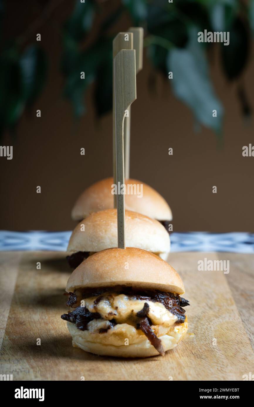 Three mini beef burgers topped with melted cheddar cheese served on a rustic wooden plank, skewered for presentation. Stock Photo