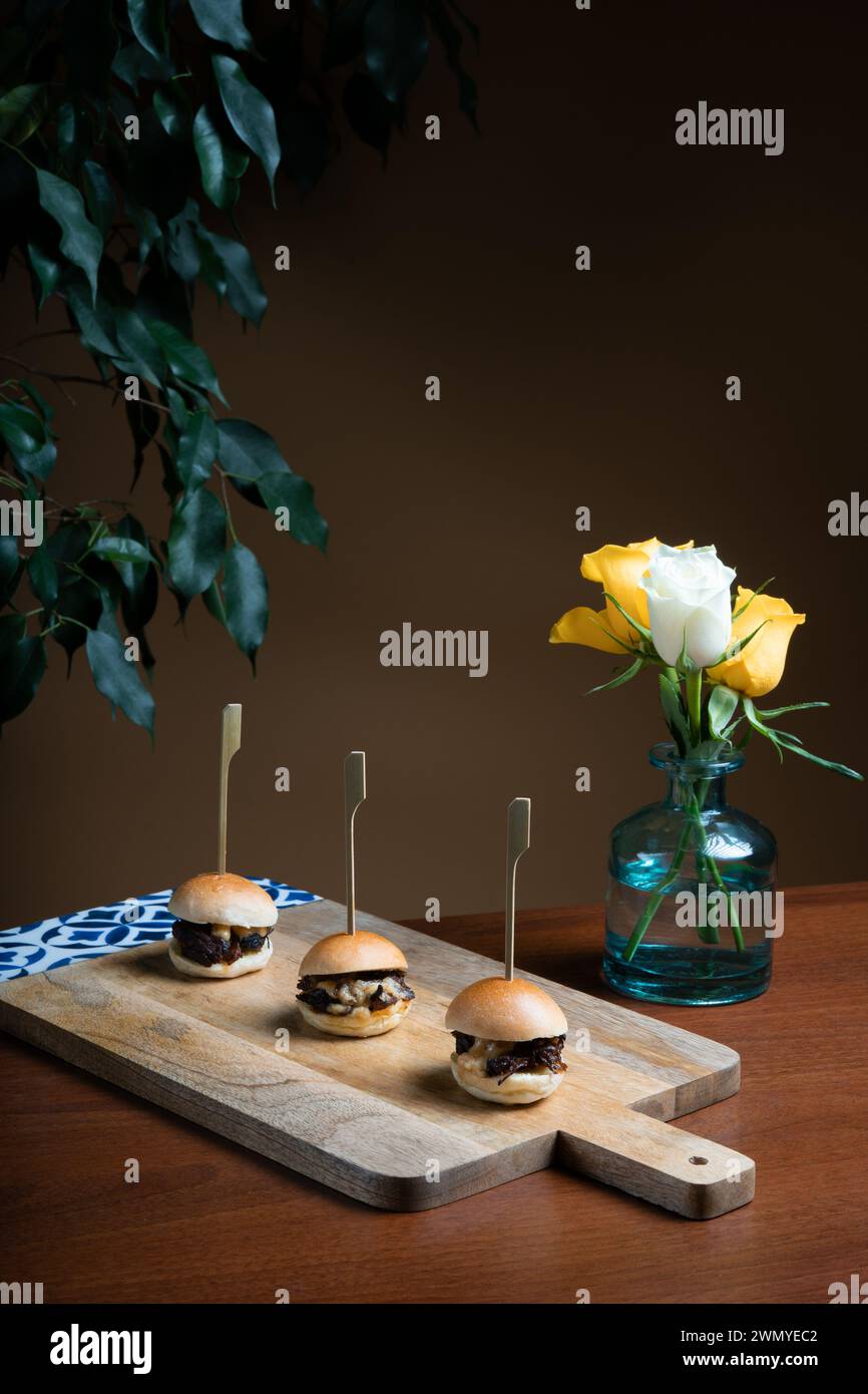 Elegant presentation of mini beef burgers topped with melted cheddar cheese, served on a wooden board with a decorative vase of roses. Stock Photo