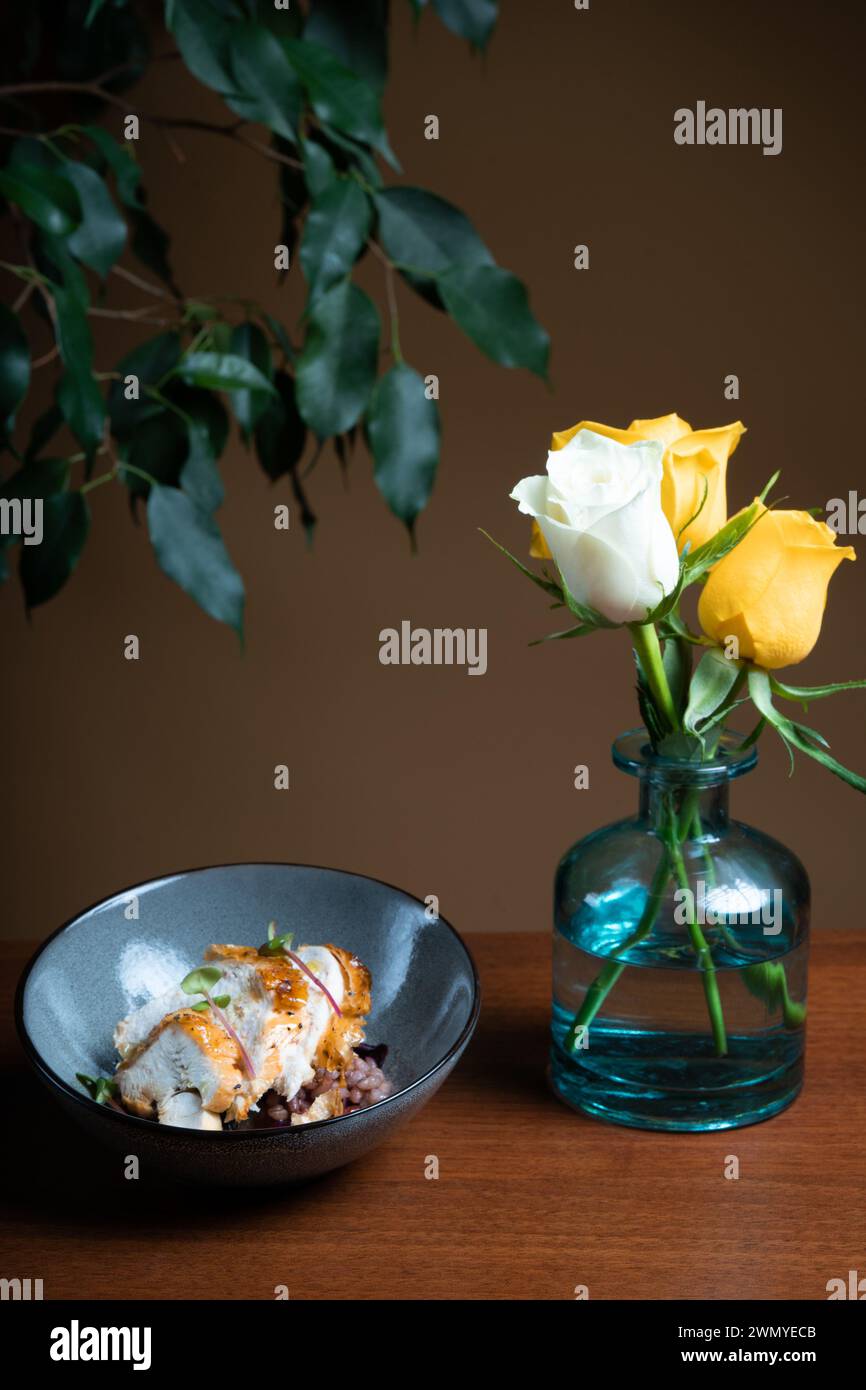 Elegant presentation of grilled chicken atop beans, garnished with herbs, on a dark plate, complemented by a glass vase of yellow roses. Stock Photo