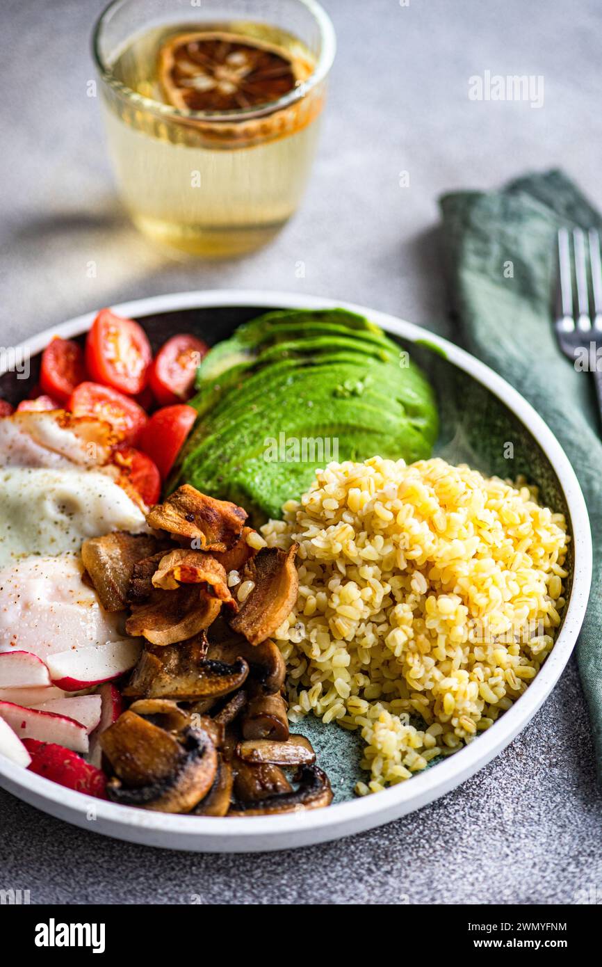Close-up of a balanced lunch with a portion of bulgur wheat, a fried egg, avocado slices, cherry tomatoes, radishes, and brown mushrooms, accompanied Stock Photo