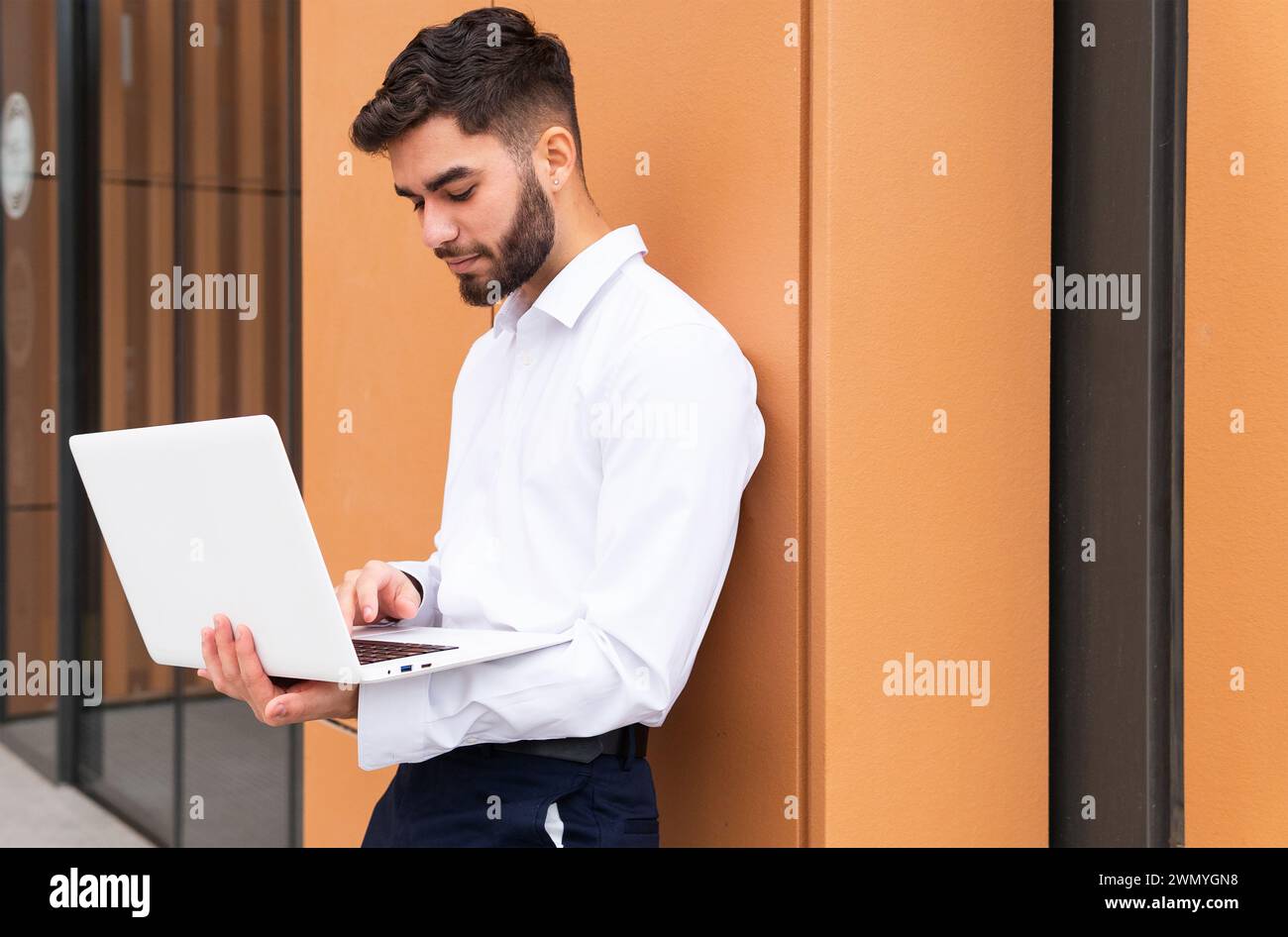 A focused young businessman in a crisp white shirt works intently on his laptop against a warm-toned, modern wall Stock Photo