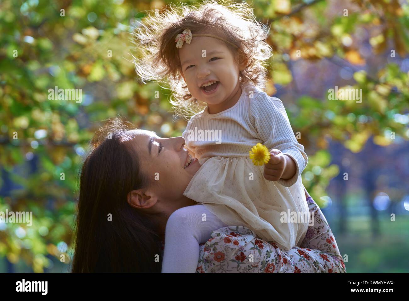 A heartwarming scene of an ethnic mother lifting her smiling daughter in a sunny Californian park, surrounded by the beauty of nature. Stock Photo