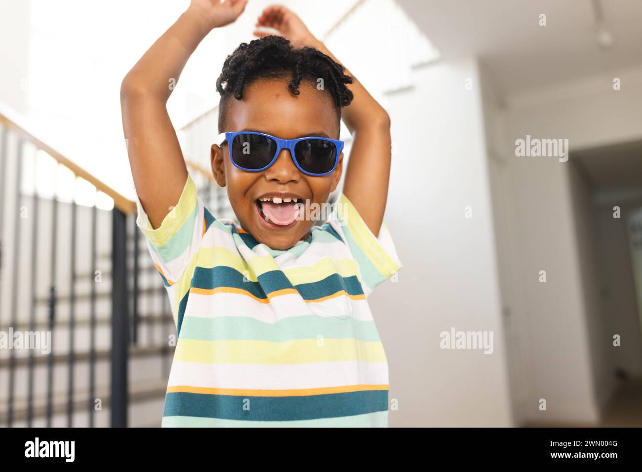 African American boy wears blue sunglasses and a striped shirt, arms raised in excitement Stock Photo