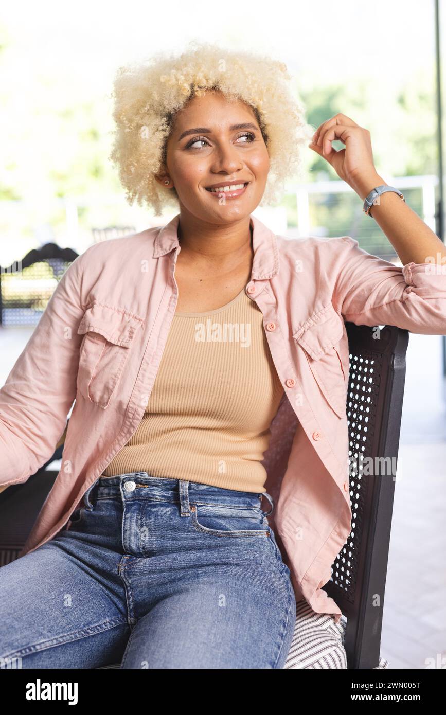 Young biracial woman with curly blonde hair sits casually in a chair Stock Photo