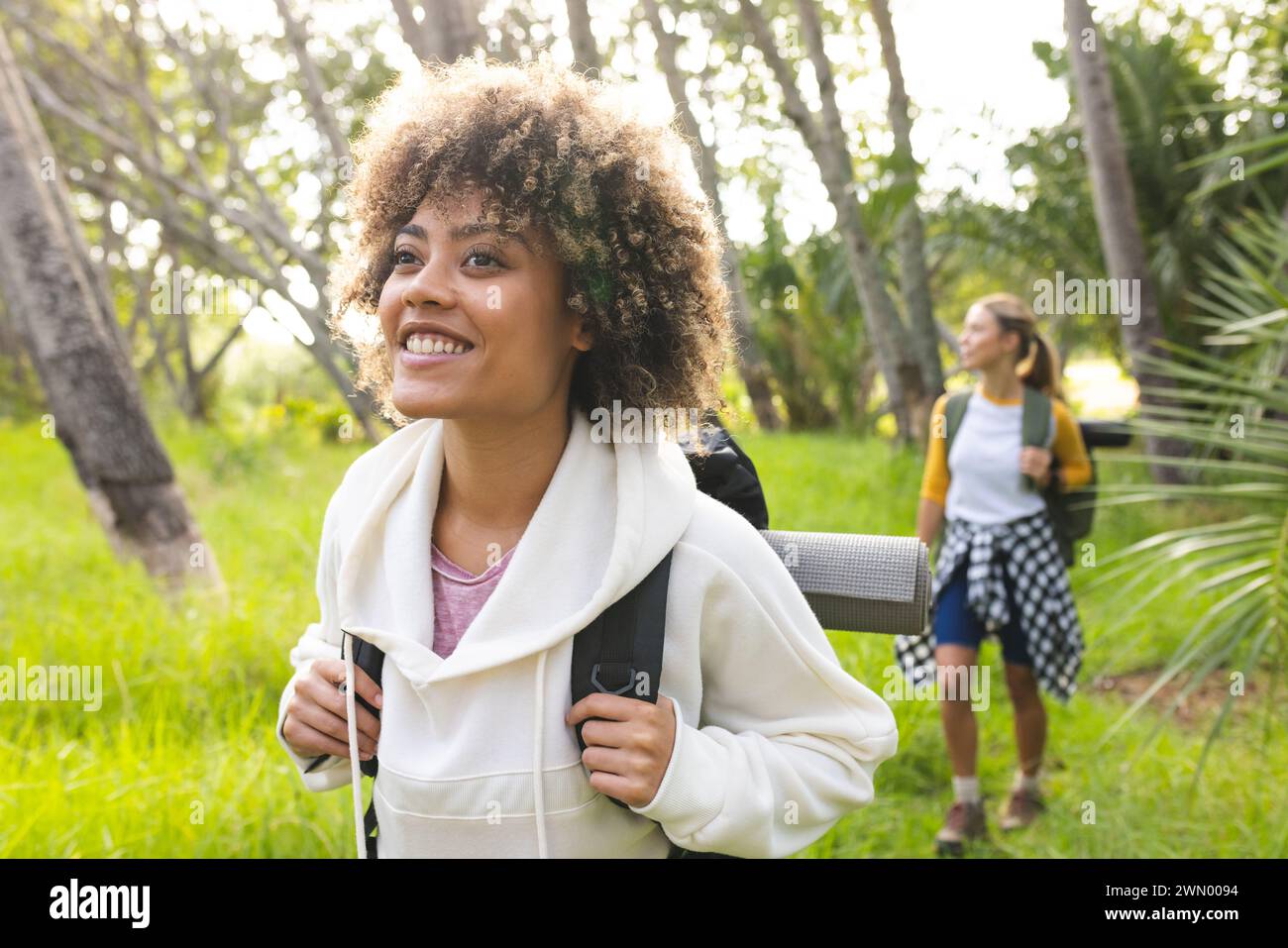 Young biracial woman with curly hair smiles while on a hike, friend in ...