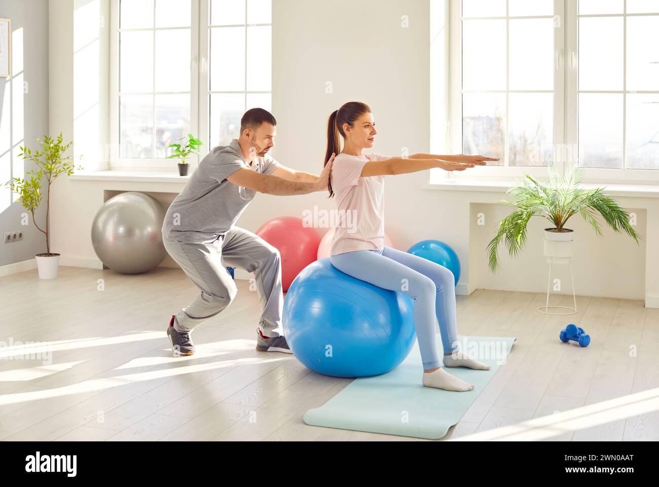 Physiotherapist helping woman doing sport exercises sitting on fit ball in rehabilitation center Stock Photo