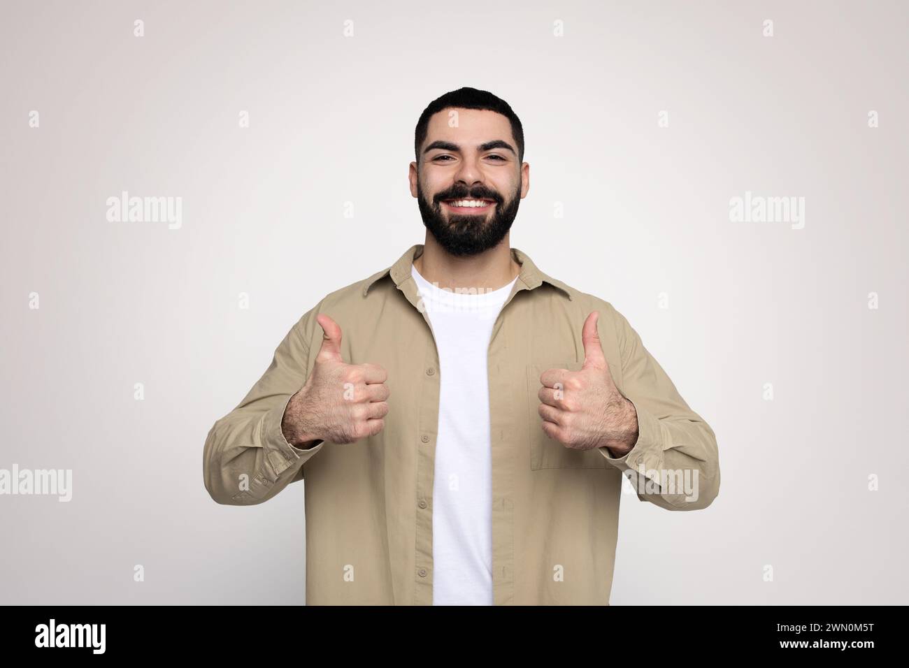 Optimistic bearded man in a beige shirt and white t-shirt giving two thumbs up with a broad smile Stock Photo