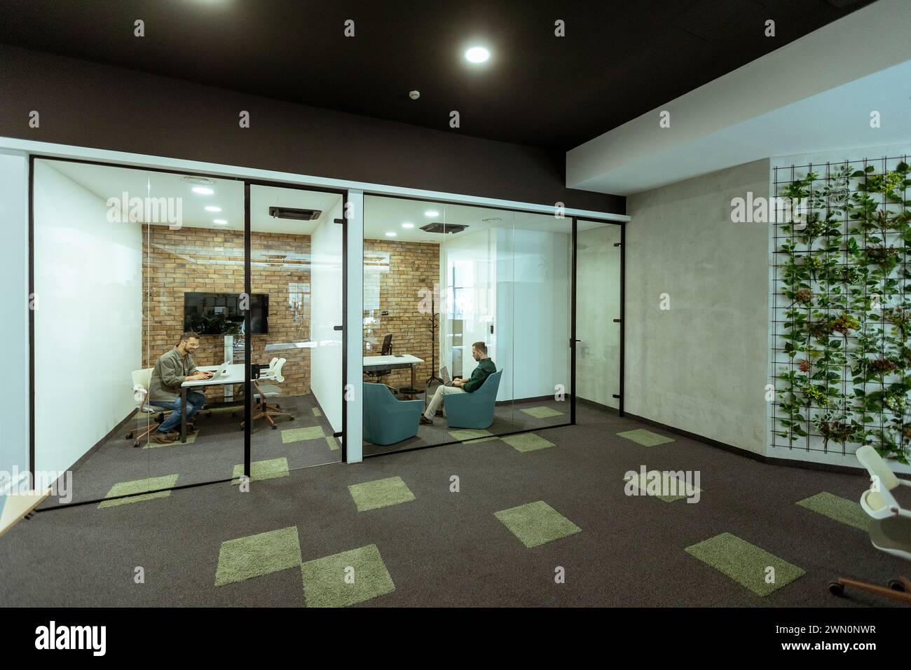 Person is immersed in a task in a stylish office with transparent glass walls. The modern design includes exposed brick, a sleek monitor, comfortable Stock Photo