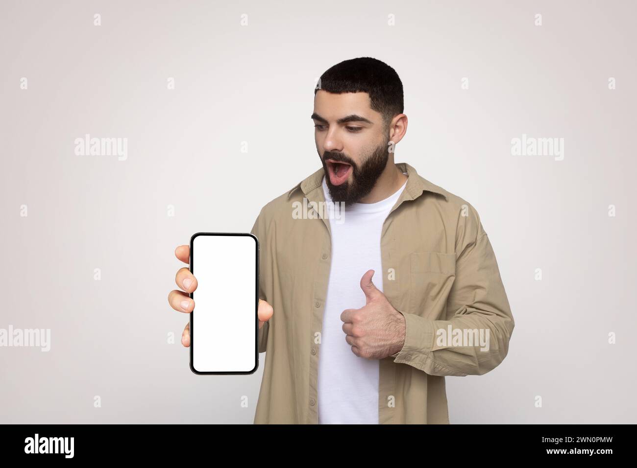 Surprised young man in a beige shirt shows a blank smartphone screen with his right hand while giving a thumbs up Stock Photo