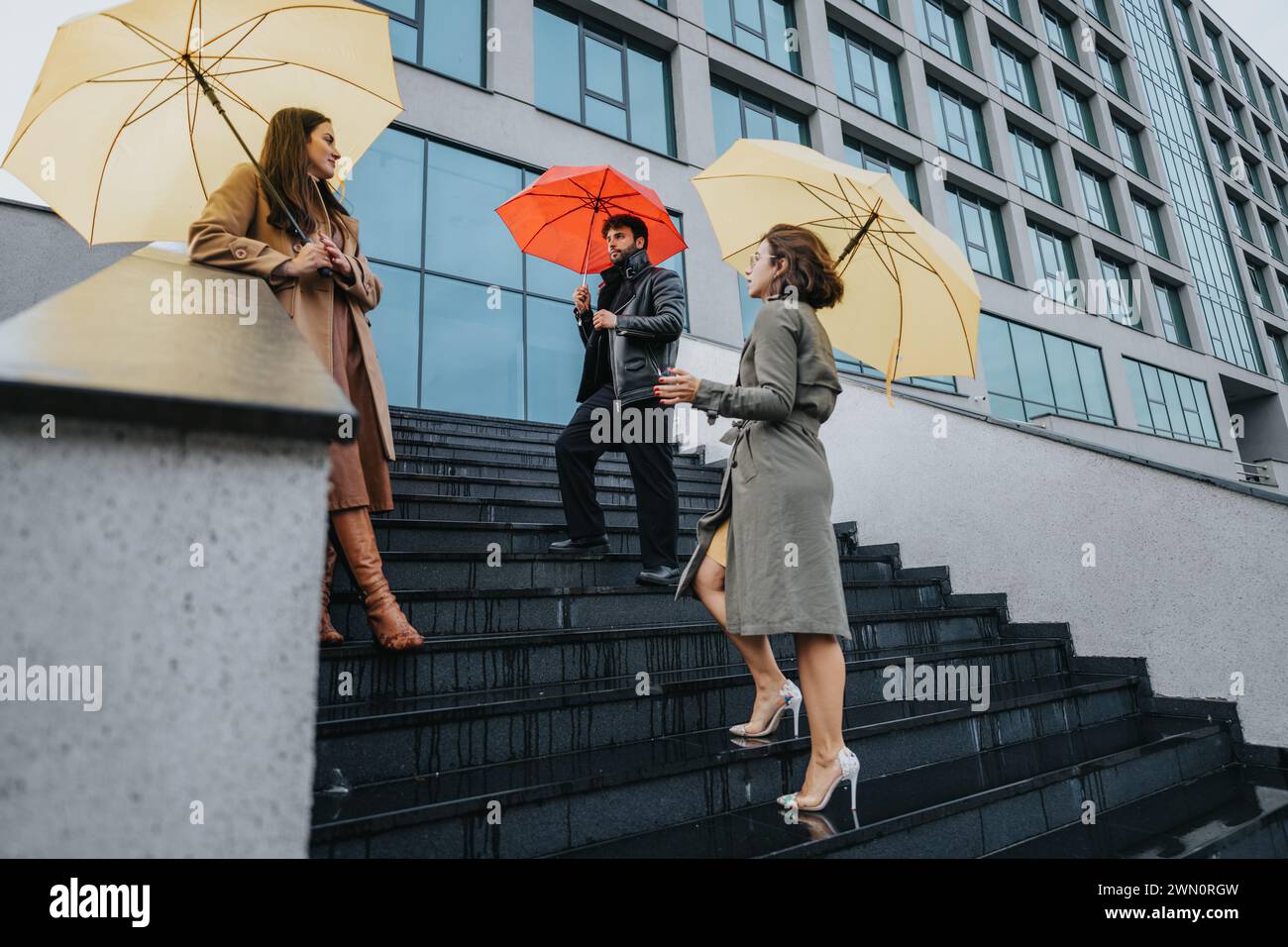 Three friends enjoy joyful moment together under vibrant yellow and red umbrellas on city steps on a rainy day, showcasing urban life and friendship. Stock Photo