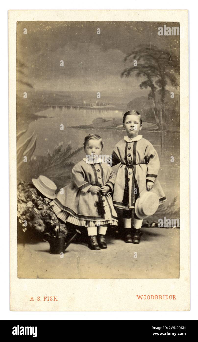 Original, charming and clear Victorian Carte de Visite (visiting card or CDV) of 2 young Victorian children, Victorian boys, siblings, brothers (not girls as have boy's hairstyles) in matching dresses with a crinoline look and pantaloons underneath for modersty holding straw hats, button detail. The older boy has a pained expression on his face. No wonder! From the photographic studio of  Alfred Samuel Fisk, John's St. Woodbridge, Suffolk, England,  U.K. circa 1865., Stock Photo