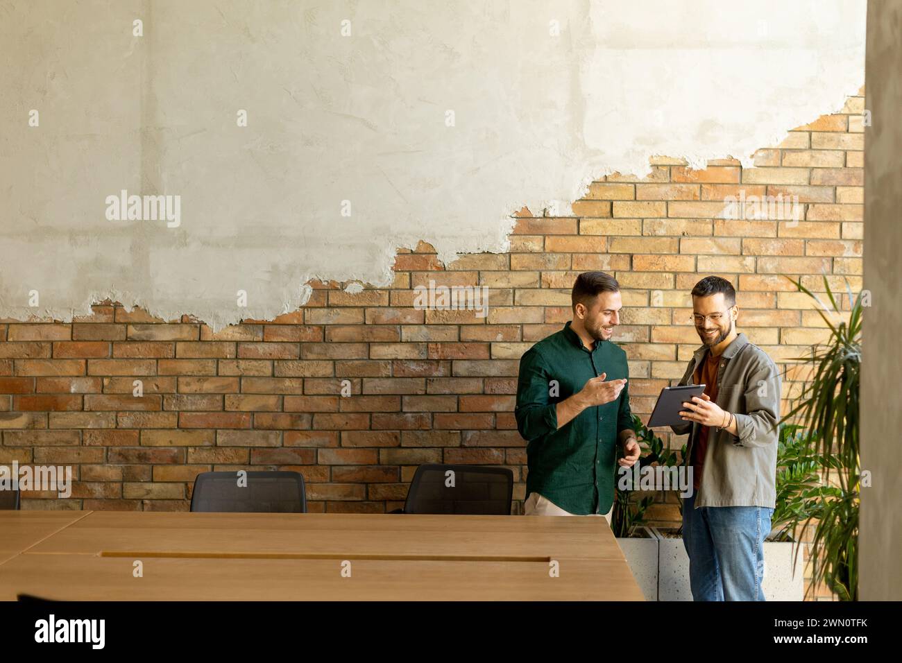 Two professionals are engaged in a focused conversation while holding digital tablet in a contemporary office space, featuring rustic exposed brickwor Stock Photo