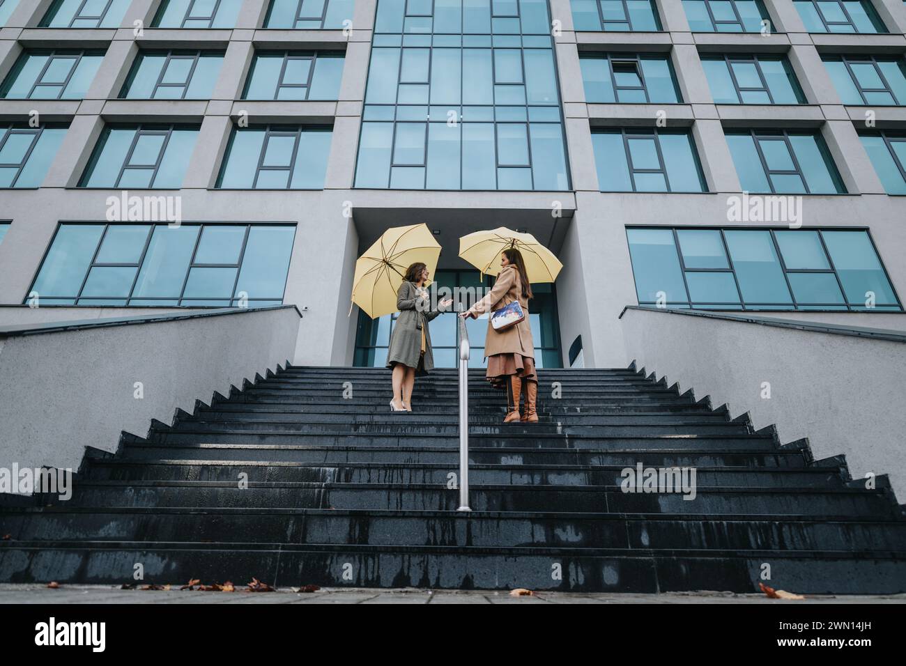Two women with yellow umbrellas on steps outside modern office building. Stock Photo