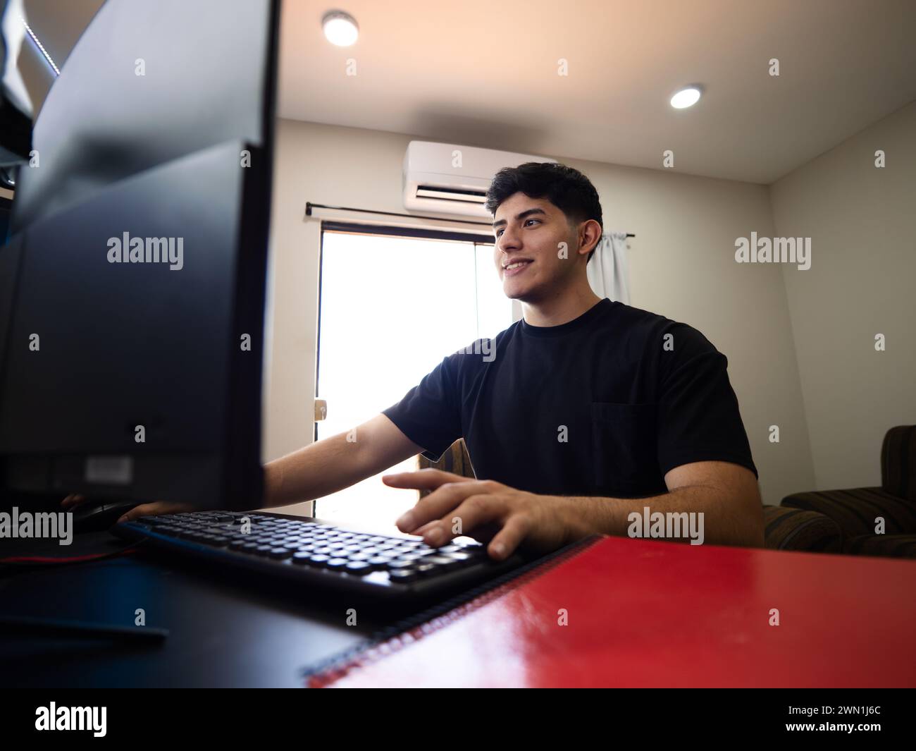 A cheerful young man stands in front of his computer, typing on the keyboard with an obvious smile Stock Photo