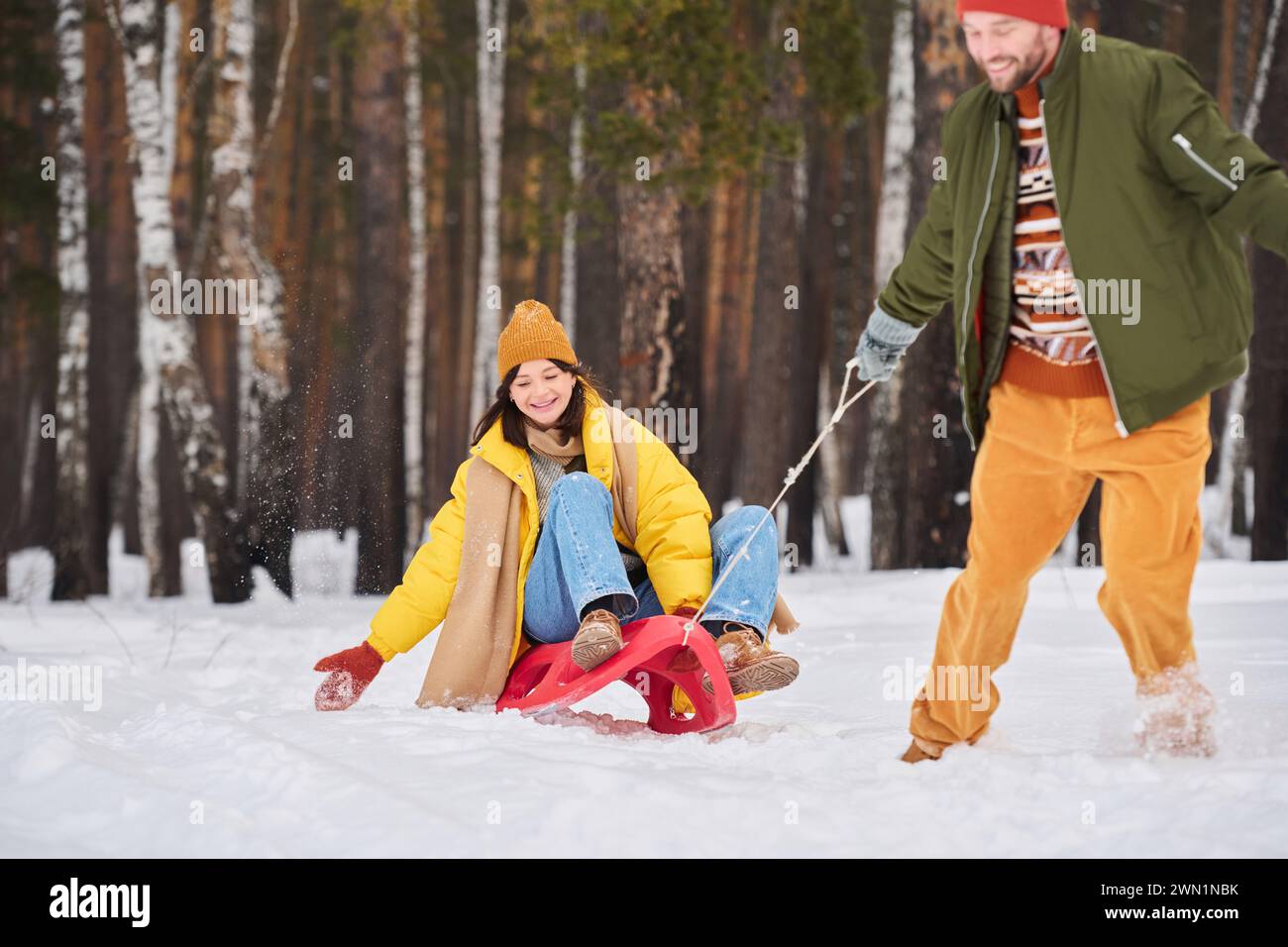 Bearded man having fun on cold winter day pulling sledge with his girlfriend sitting on it Stock Photo