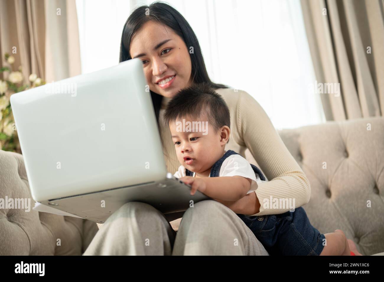 A happy Asian businesswoman mom is working from home, working on her laptop, and taking care of her naughty baby boy. Stock Photo