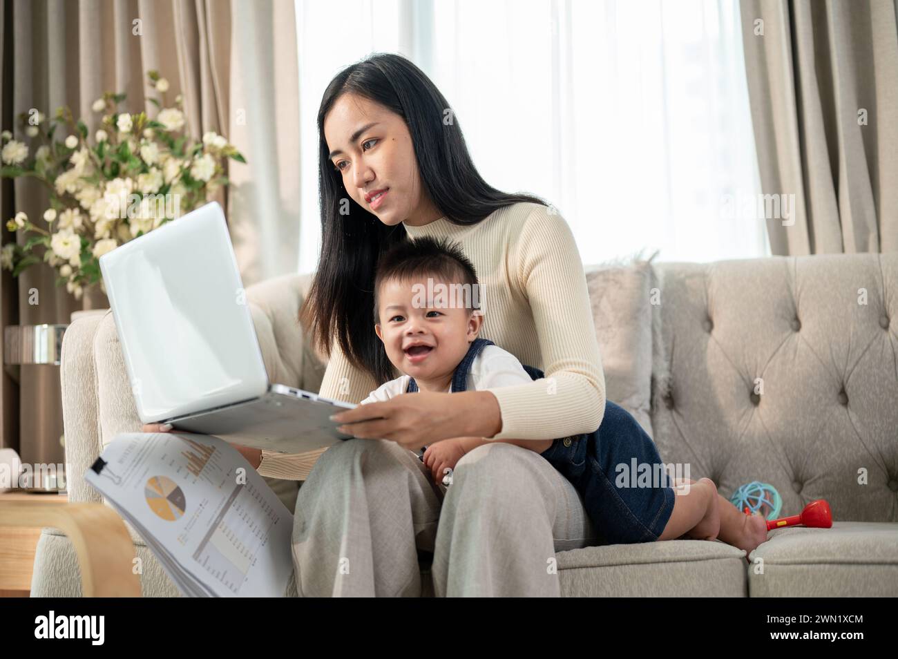 A happy Asian businesswoman mom is working from home, working on her laptop, and taking care of her naughty baby boy. Stock Photo