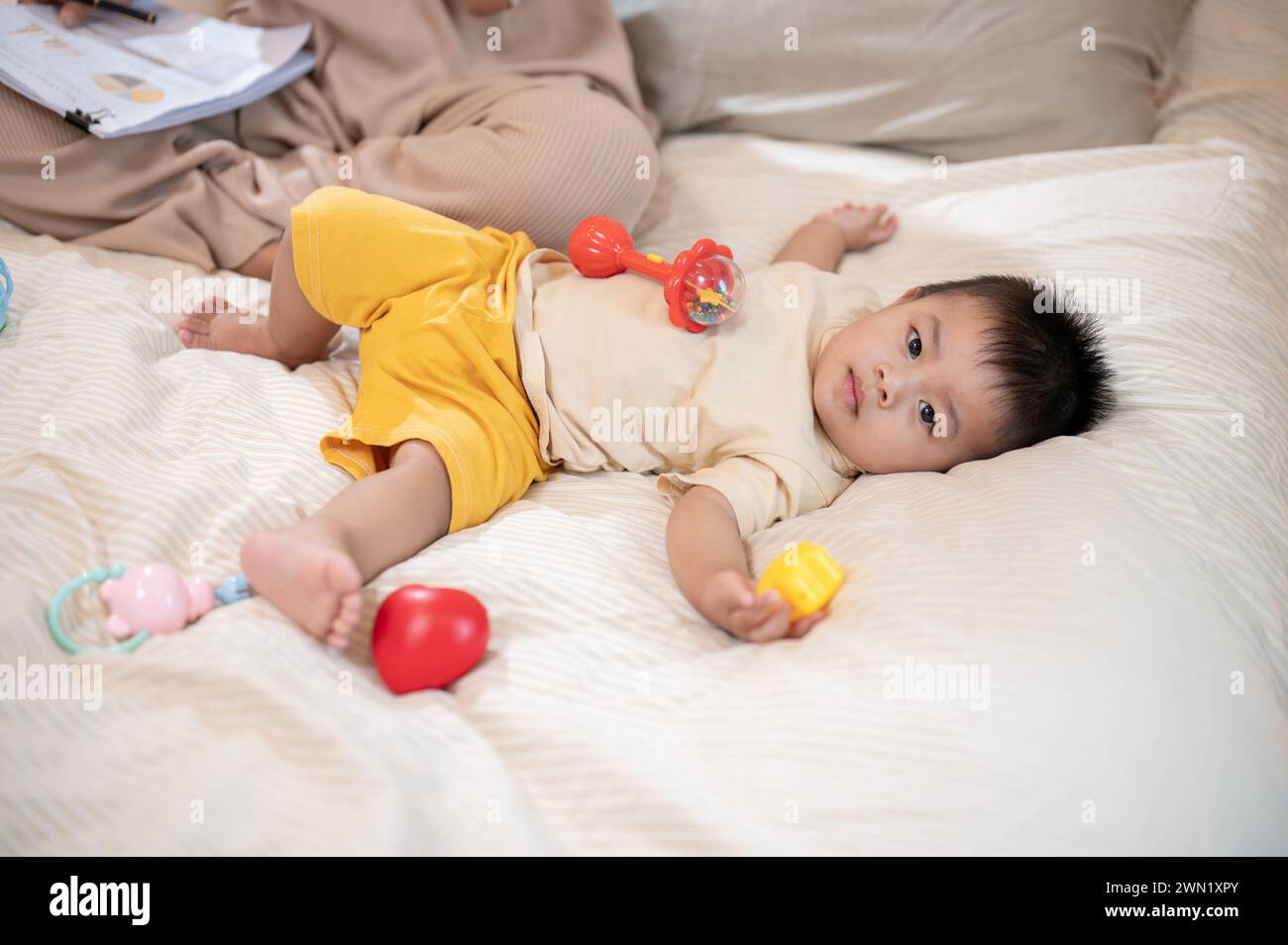 A cute, innocent Asian baby boy is playing with toys while lying on the bed in the bedroom. toddler, little boy, son, child Stock Photo