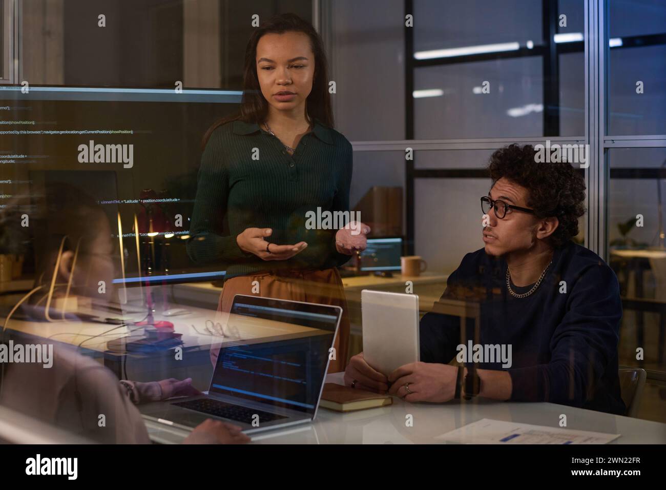 Young female programmer standing at table having discussion with her colleagues in modern office in evening Stock Photo