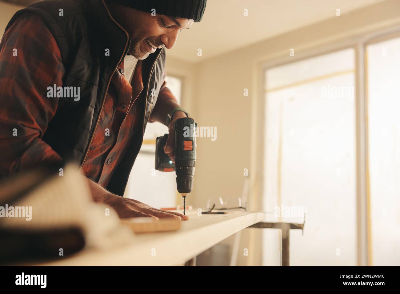 Happy man using a power drill to secure a plank for baseboards in a home interior. Mature man renovating and refurbishing his kitchen using his self-t Stock Photo